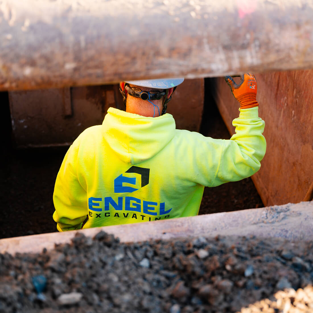 Worker in neon Engel hoodie, hard hat, in dirt trench.