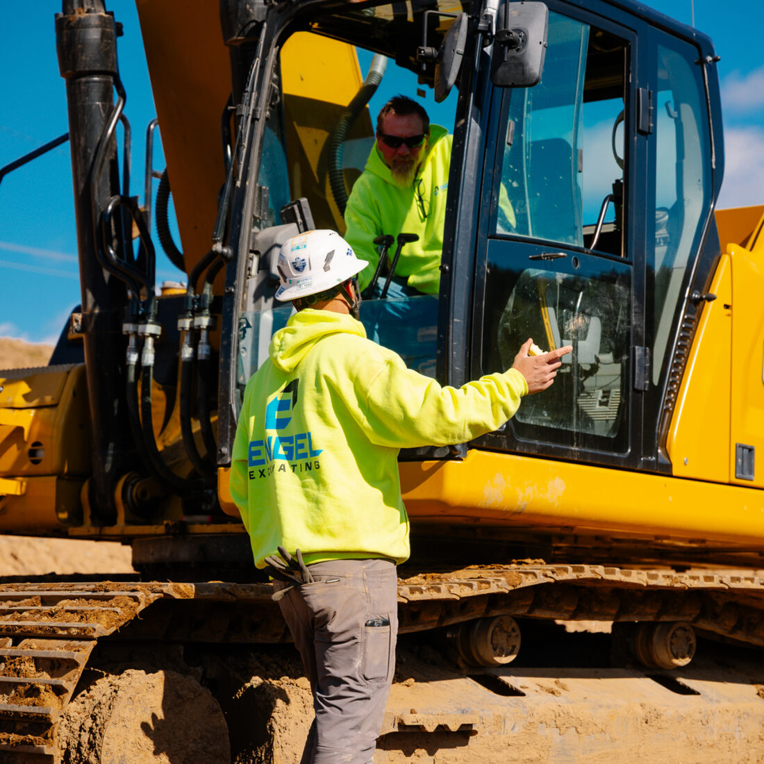 Two workers in yellow hoodies by an excavator.