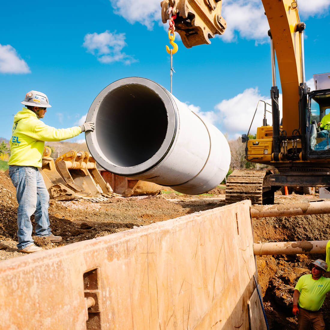 Workers guide concrete pipe lifted by excavator outdoors.