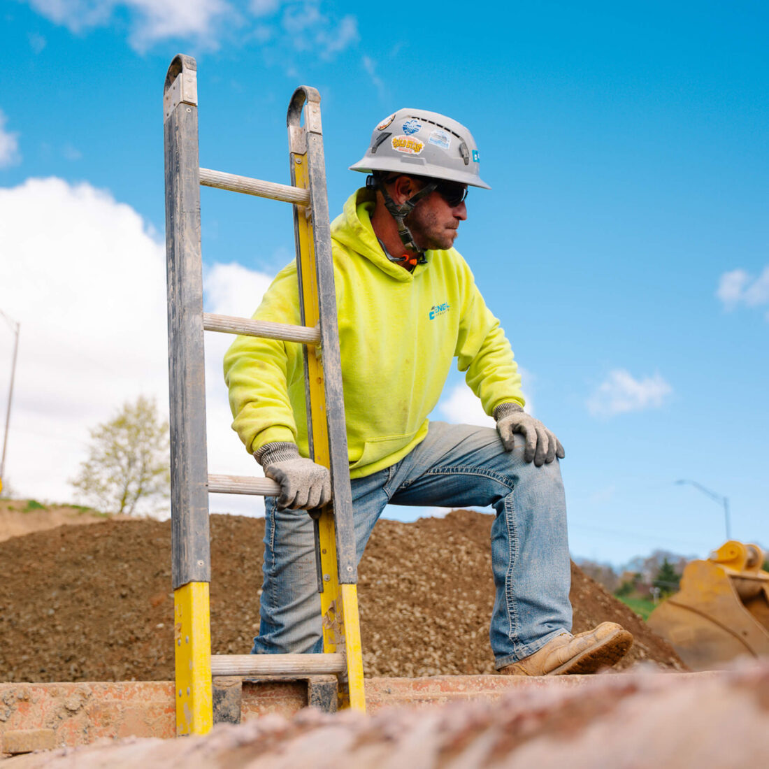 Worker in yellow hoodie, helmet by ladder on dirt site.