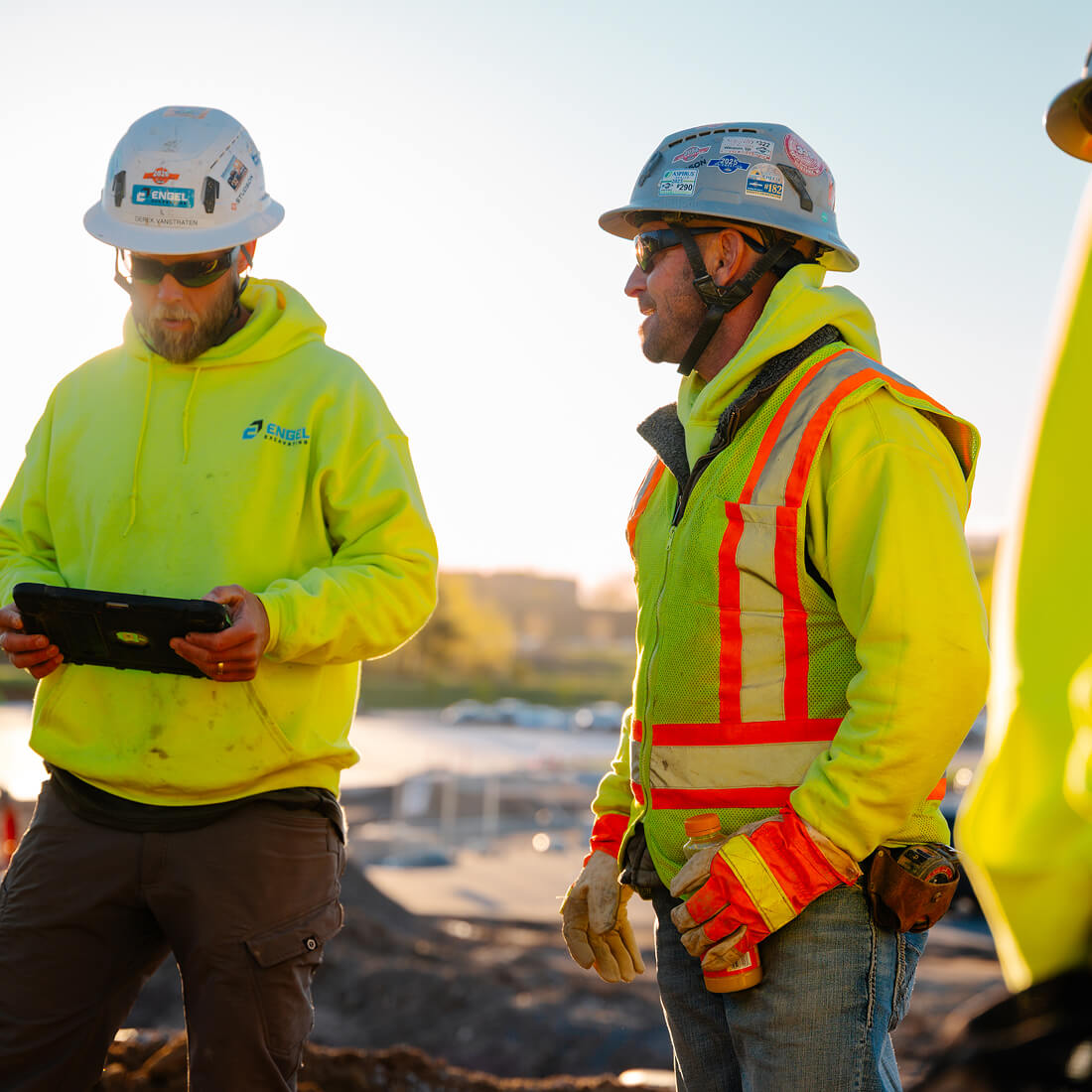 Construction workers in safety gear, one holds tablet, outdoors.