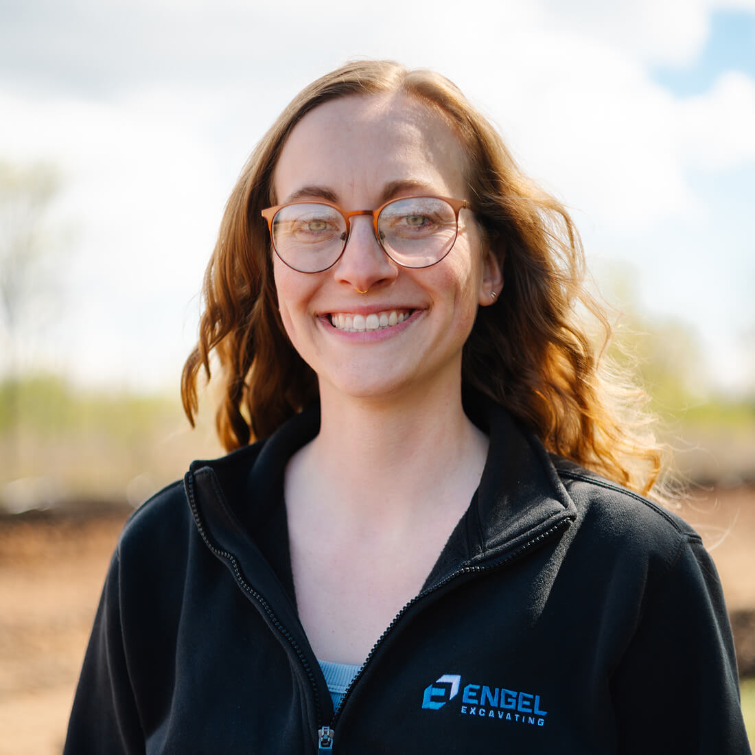 Woman in glasses smiling outdoors, blurred trees behind her.