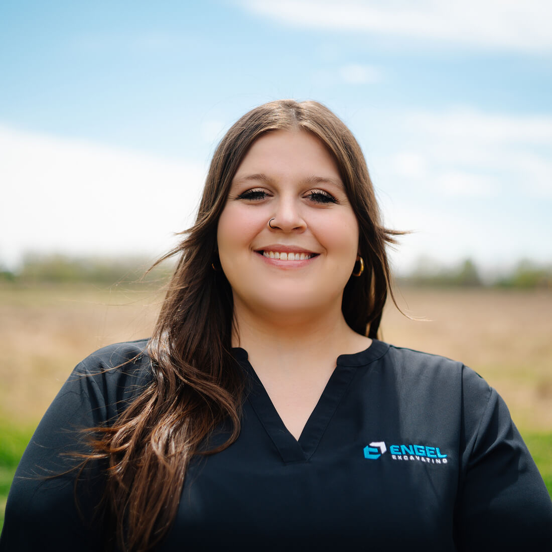 Smiling woman in black shirt outdoors, field and blue sky.