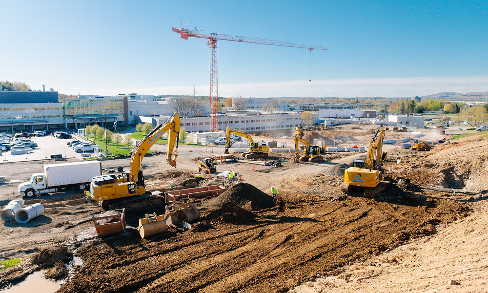 Construction site with excavators, workers, trucks, crane, buildings.