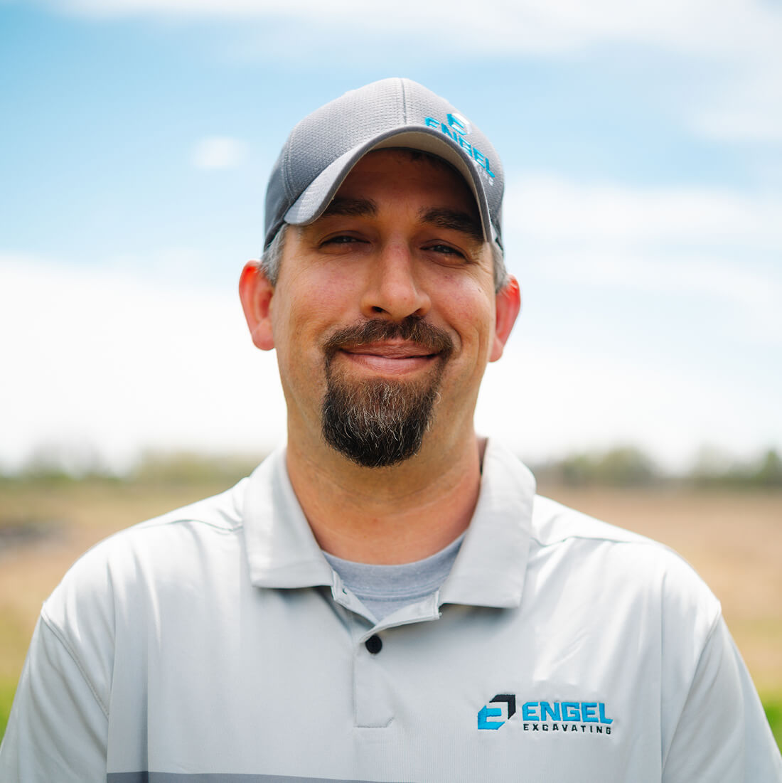 Man in gray Engel Excavating polo and cap outdoors, smiling.