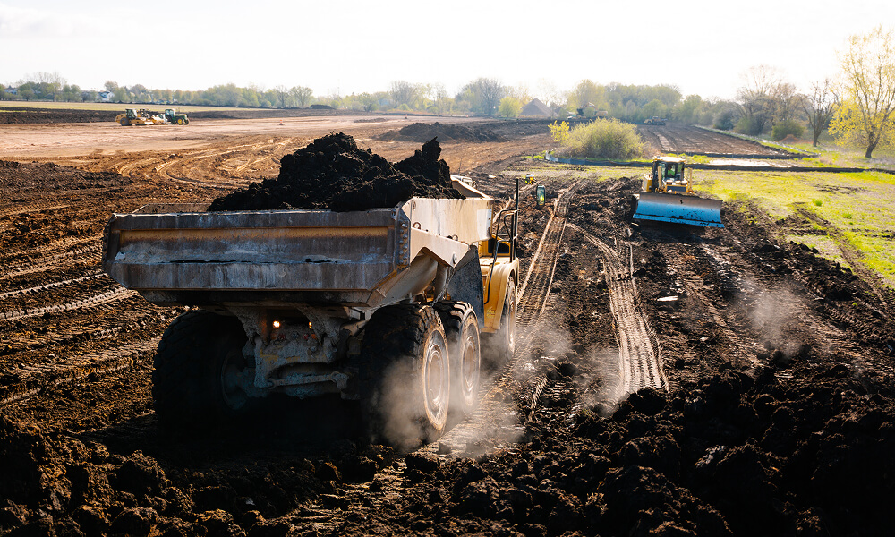 Dump truck drives on muddy site, machine levels ground nearby.