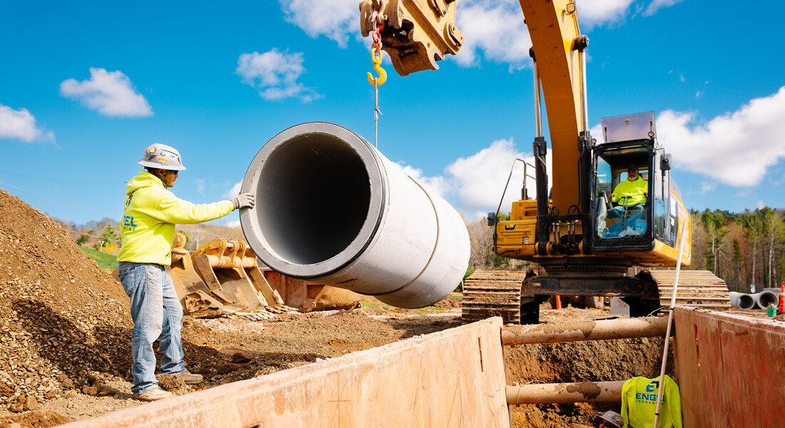 Worker guides concrete pipe lifted by excavator on site.
