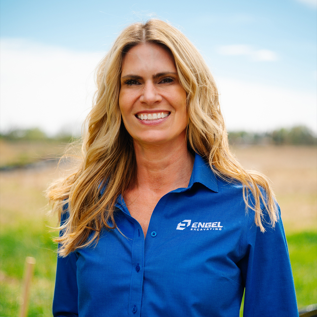 Smiling blonde woman in blue shirt stands outside on grass.