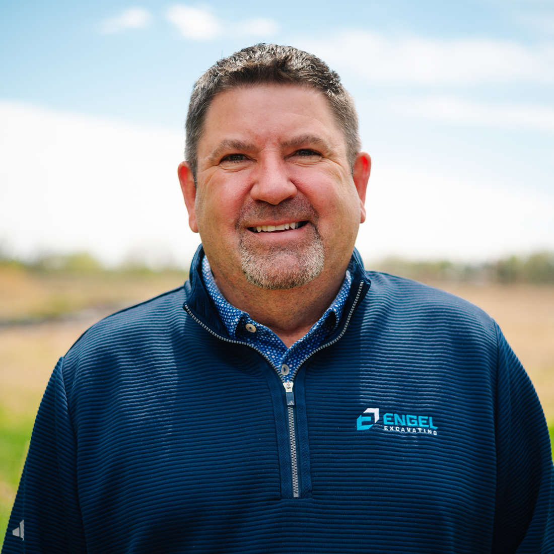 Man in dark blue pullover smiles outdoors in sunlight.