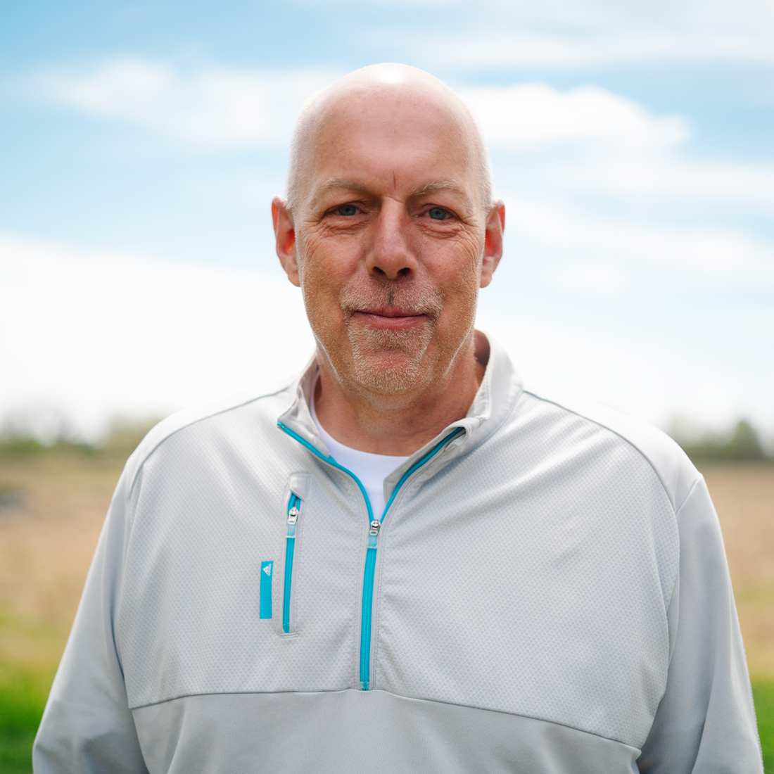 Man in gray jacket outdoors, field and trees behind.